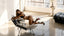 Woman lounging in a black lingerie seton a chair in a modern, sunlit room with large windows.